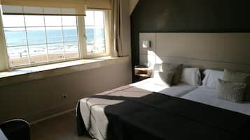 Habitación de hotel con una cama grande, una mesita de noche con lámpara y vista a la playa a través de una ventana.