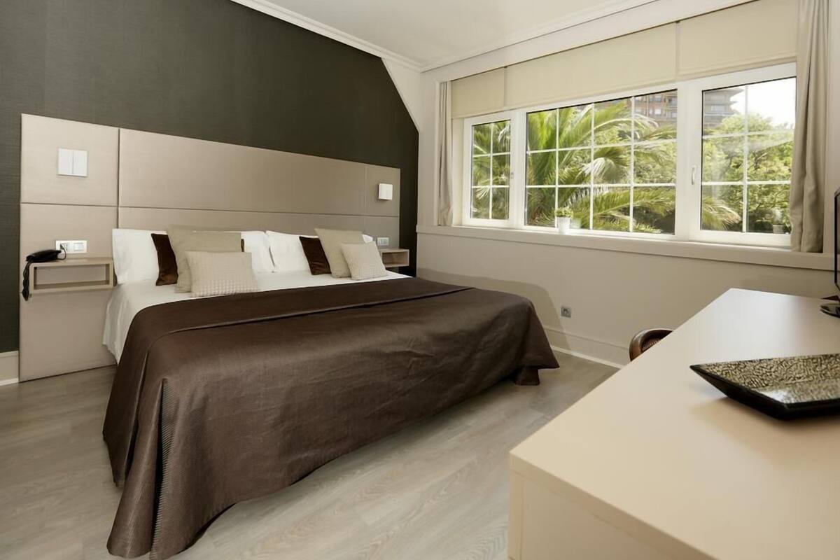 A bedroom with a large bed, a nightstand, a desk, and a window with a view of greenery.