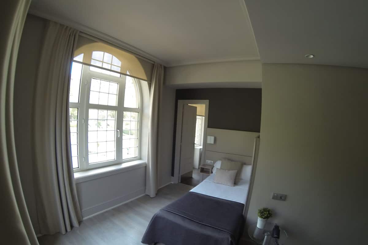 A bedroom with a large window, a bed, a bedside table, and a small potted plant.