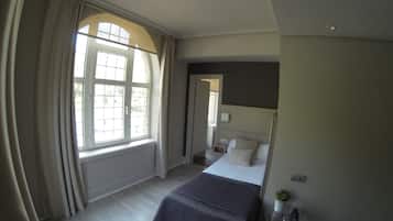 A bedroom with a large window, a bed, a bedside table, and a small potted plant.
