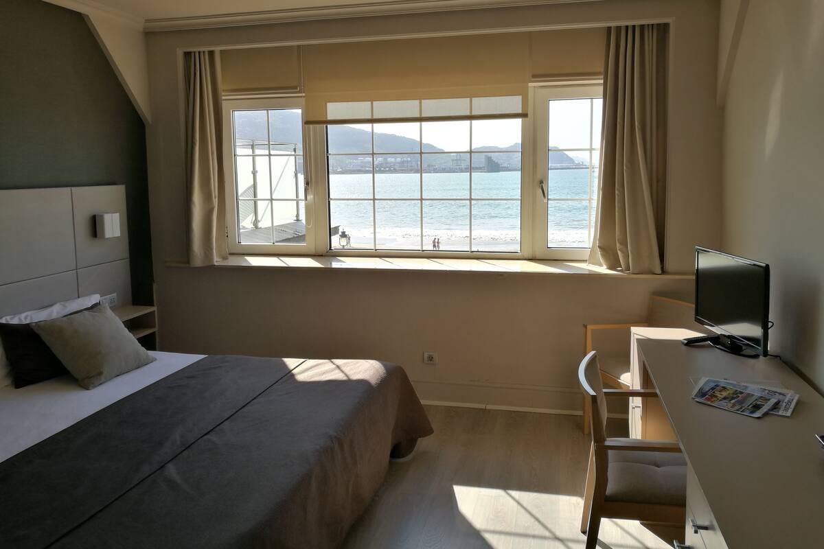 A hotel room with a bed, a desk, and a large window with a view of the sea.