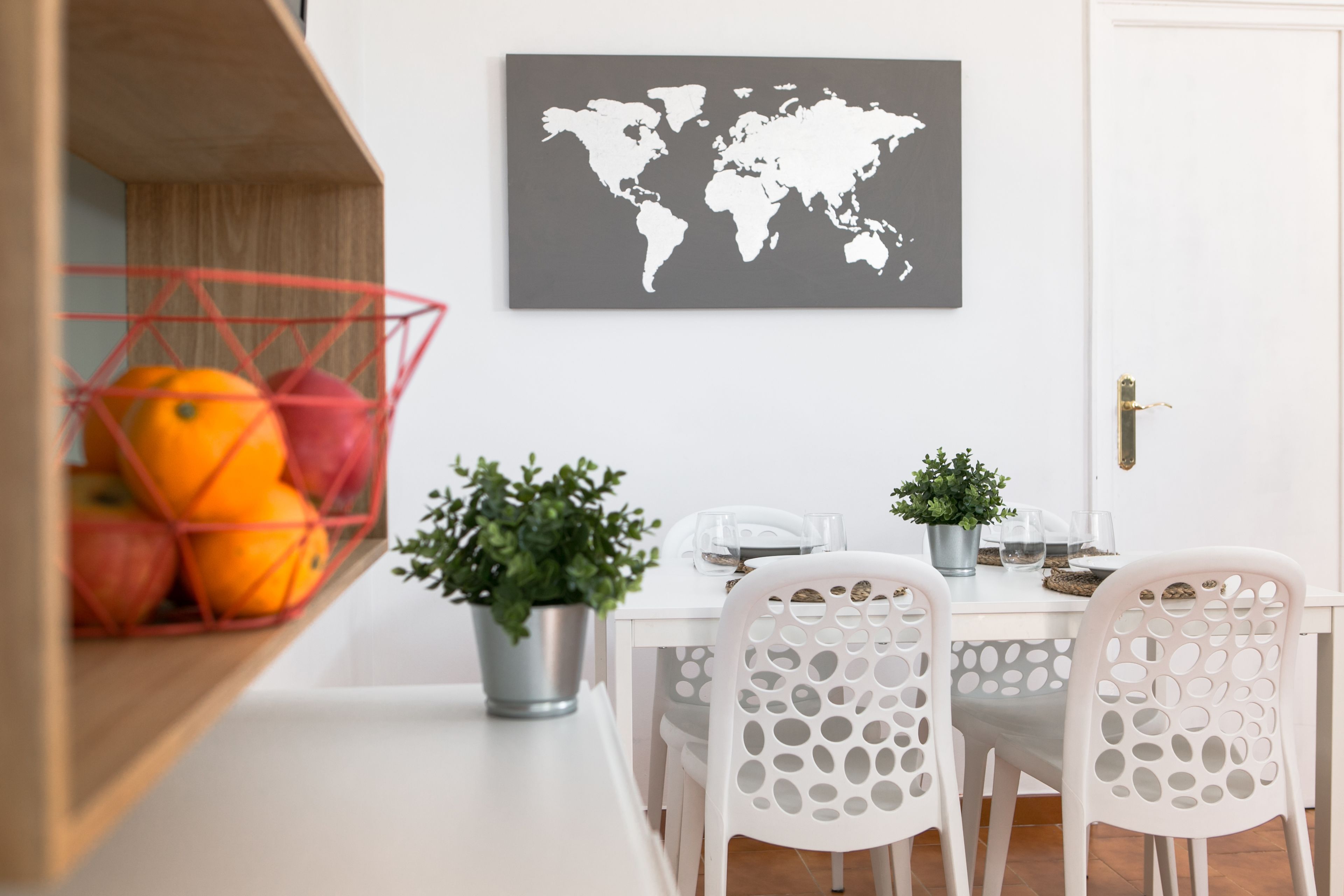 A dining area with a white table, four white chairs with circular cutouts, a world map on the wall, a metal fruit basket with oranges and apples, and a potted plant on the table.
