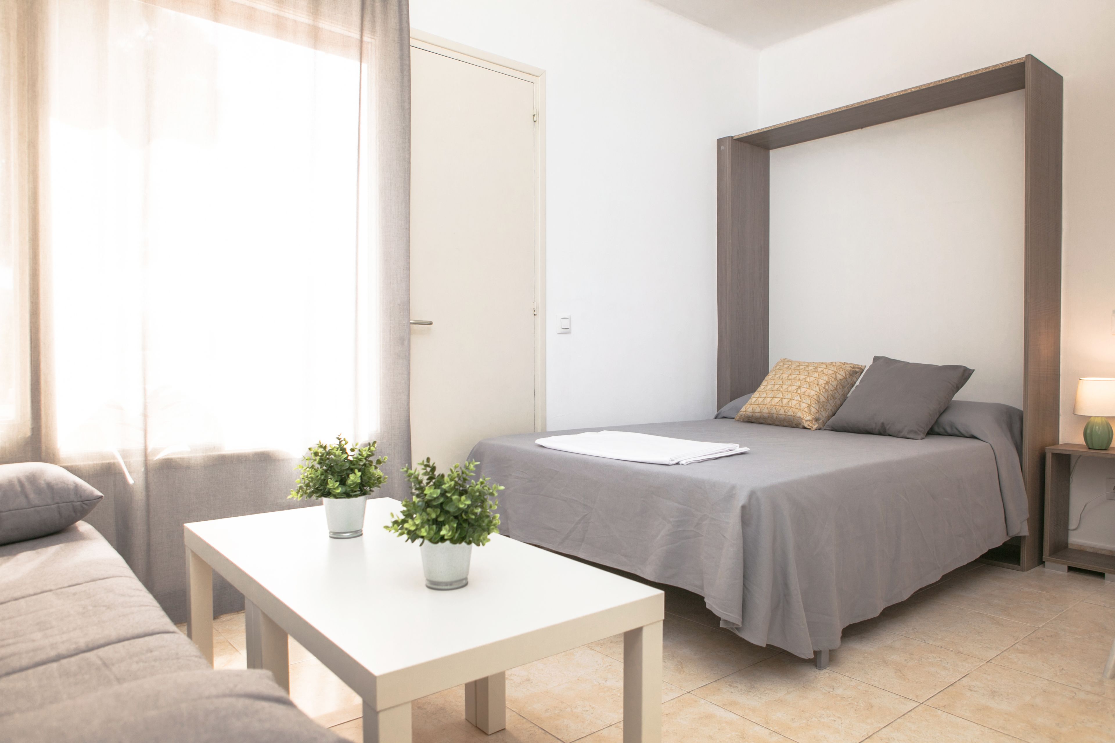 Un dormitorio moderno con una cama, una mesita con plantas, un sofá y una mesita de noche con una lámpara.