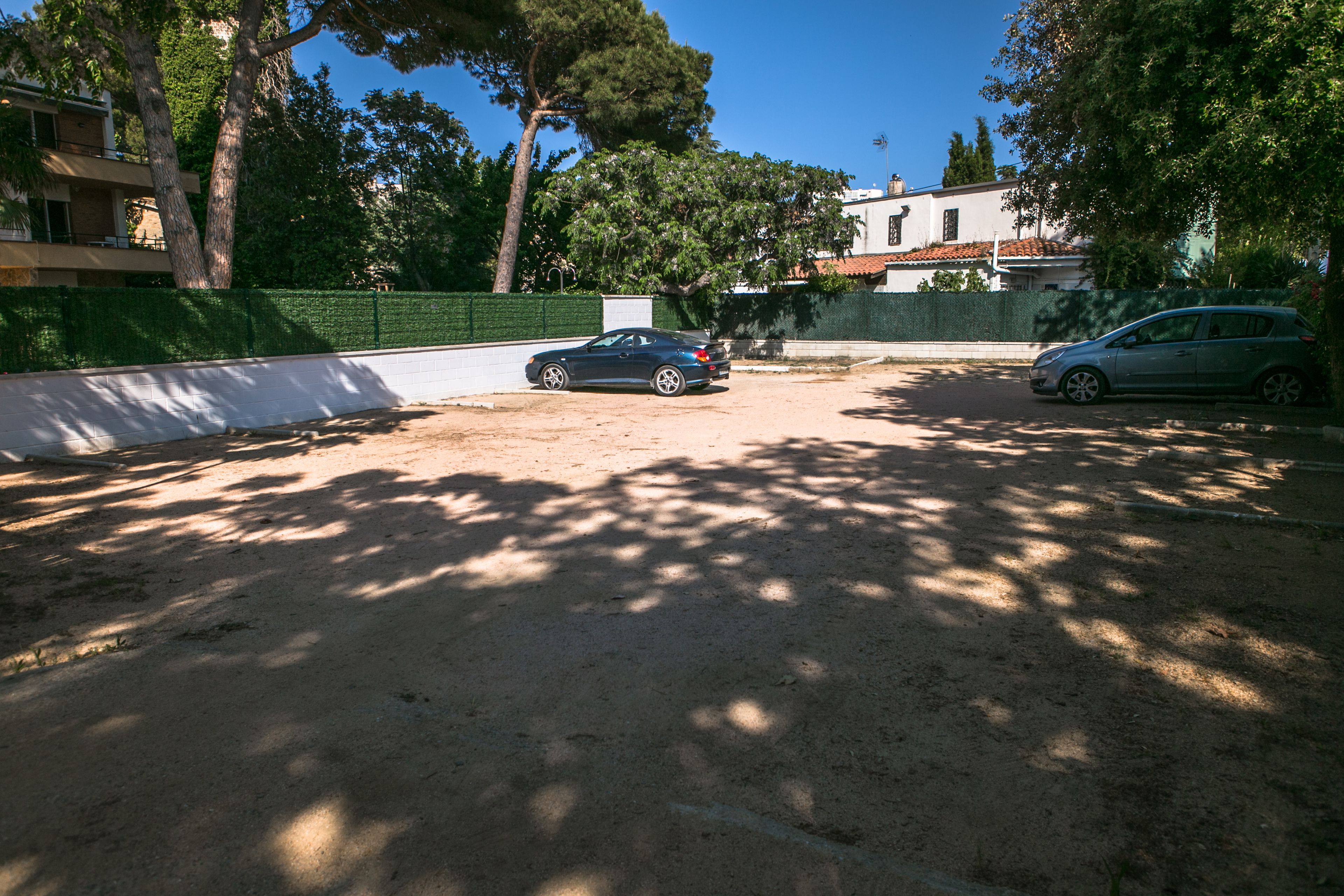 A parking area with two cars, surrounded by trees and a green fence.