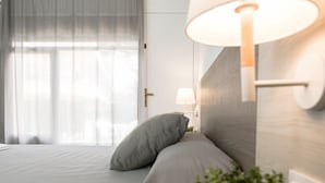 A neatly made bed with a pillow, a bedside lamp, and a window with sheer curtains. - RVApartaments Benelux (Castell-Platja d'Aro)