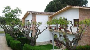A two-story building with a white facade and a brown roof. There are trees in front of the building, and a paved path leading to the entrance. - RVApartaments Benelux (Castell-Platja d'Aro)