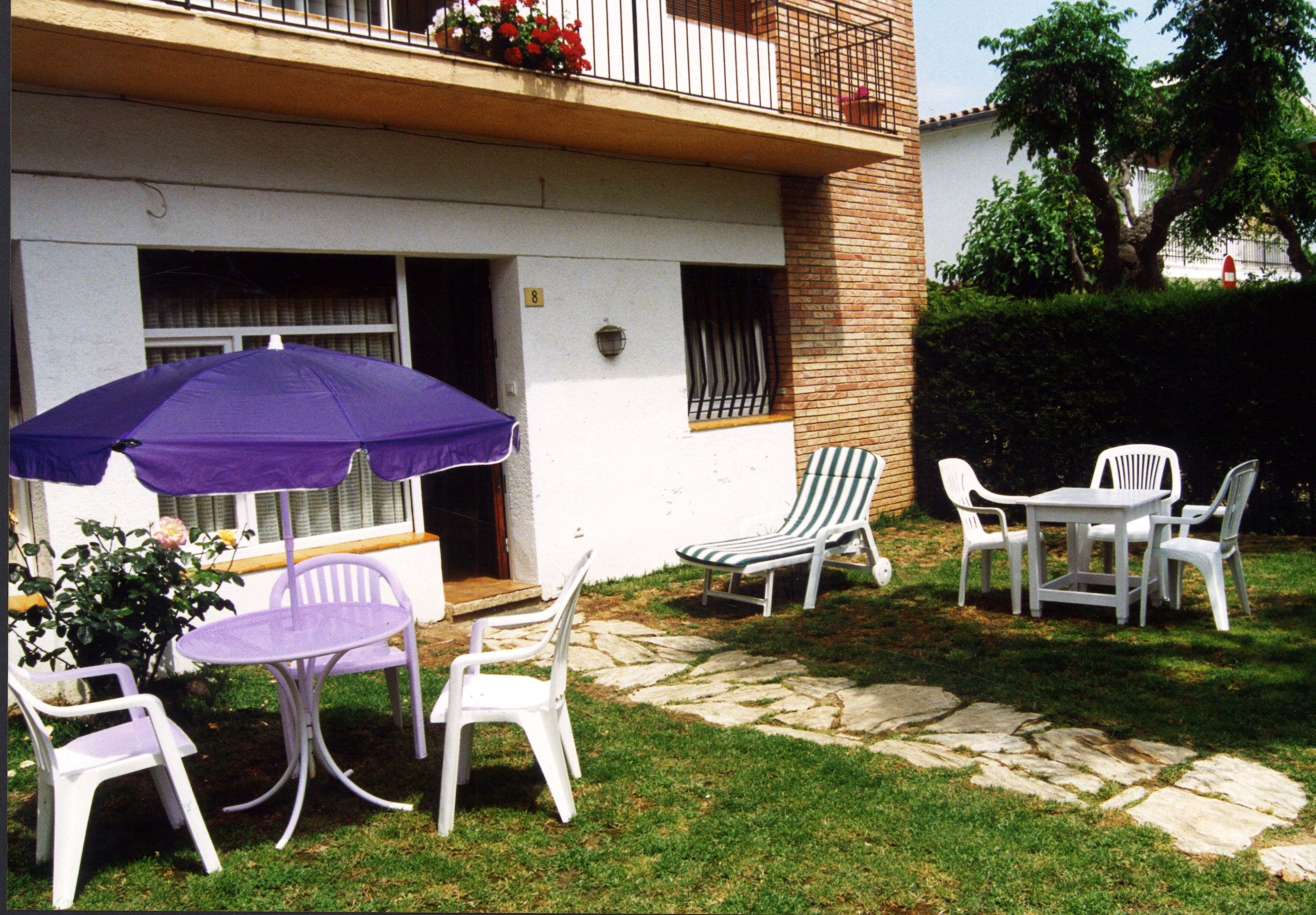 Un pati exterior amb caires de plàstic blanc, una taula, un parasol morat i un camí de pedra.