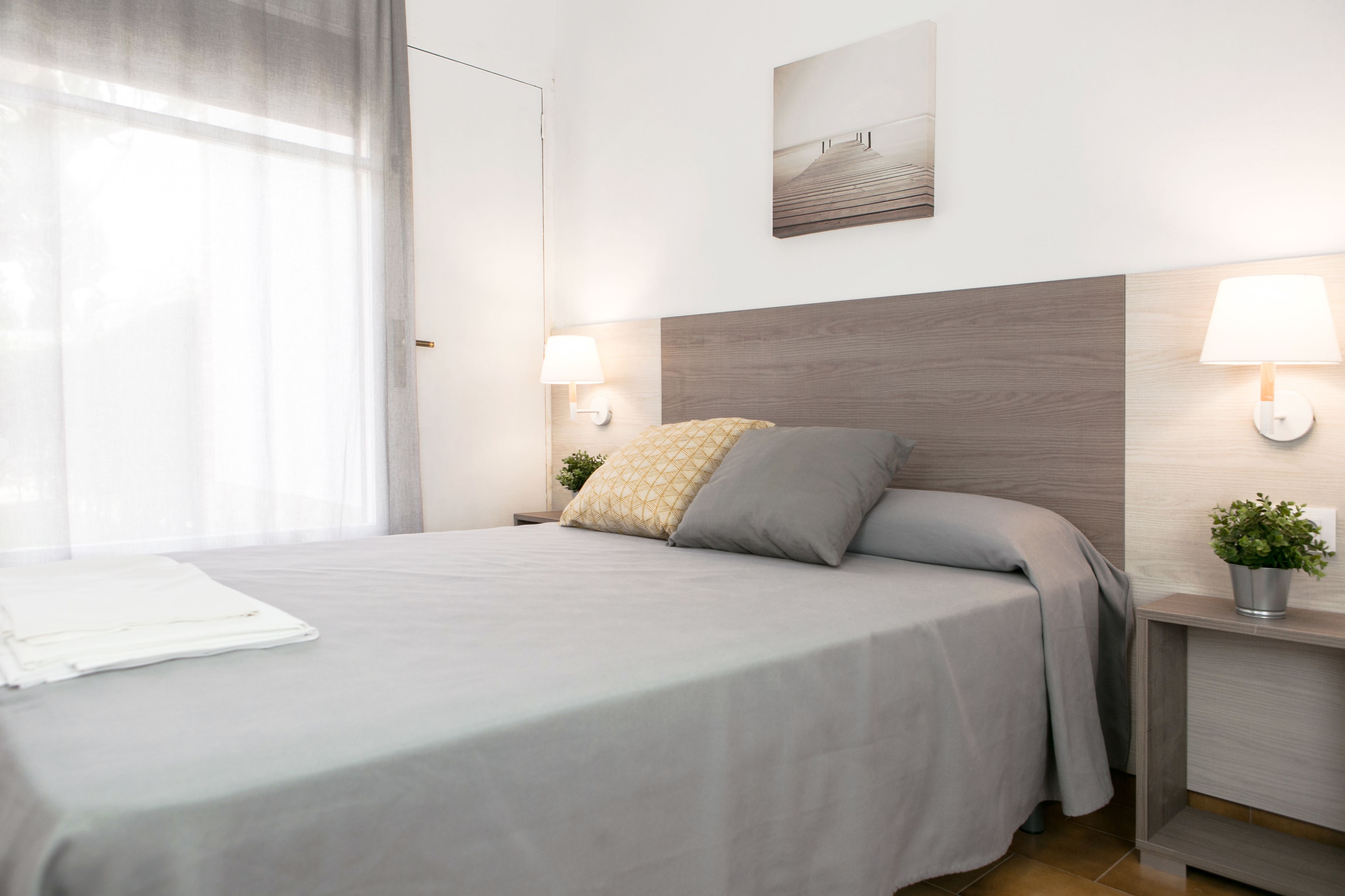 Una cama bien hecha con una colcha gris y dos almohadas, una mesita de noche con una lámpara, un cuadro en la pared y una ventana con cortinas translúcidas.
