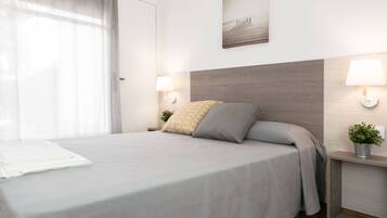 Una cama bien hecha con una colcha gris y dos almohadas, una mesita de noche con una lámpara, un cuadro en la pared y una ventana con cortinas translúcidas.