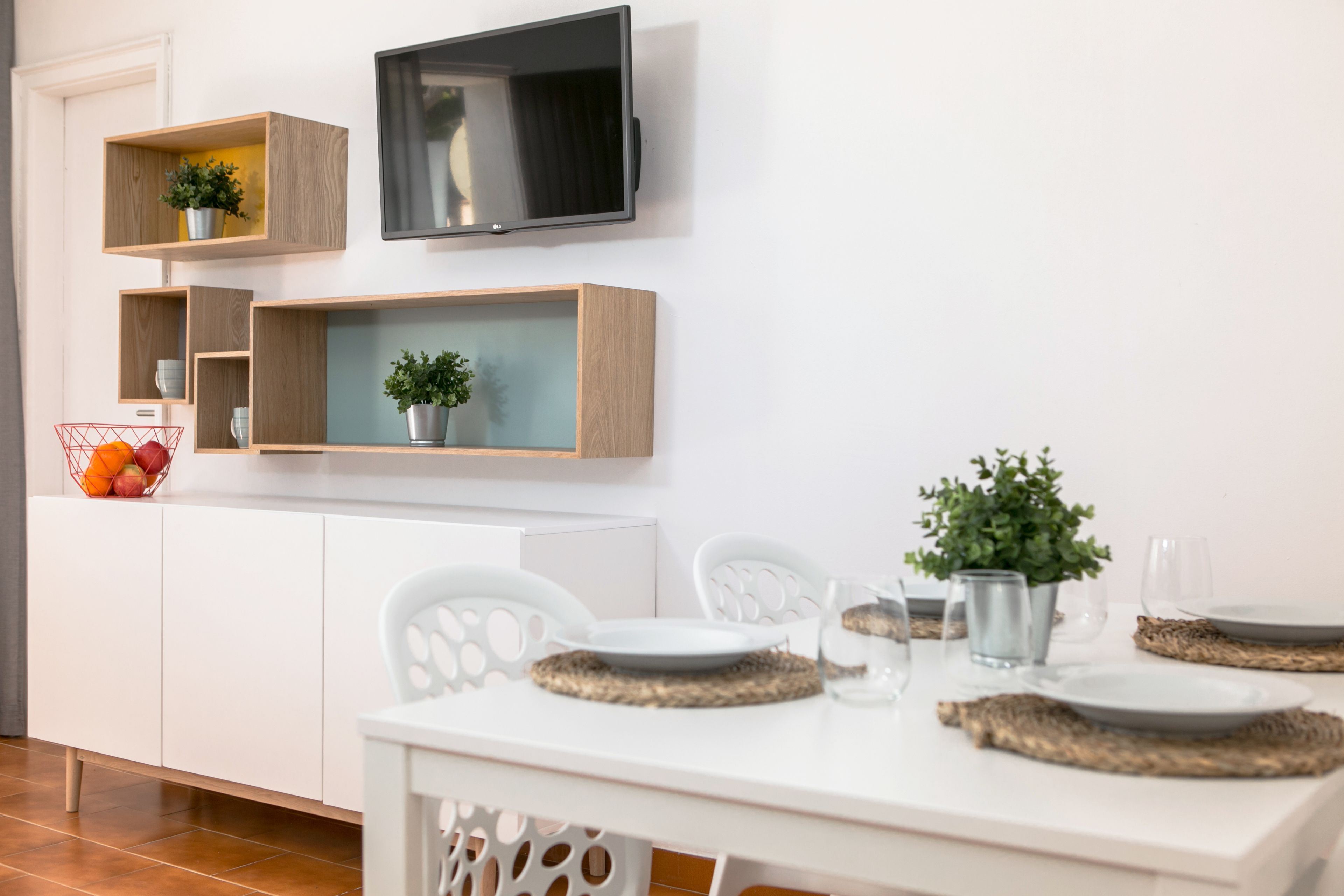 A modern dining area with a white table set for four, surrounded by white chairs. There are two wooden shelves with plants and a basket of fruit on the left. A flat-screen TV is mounted on the wall above the table.