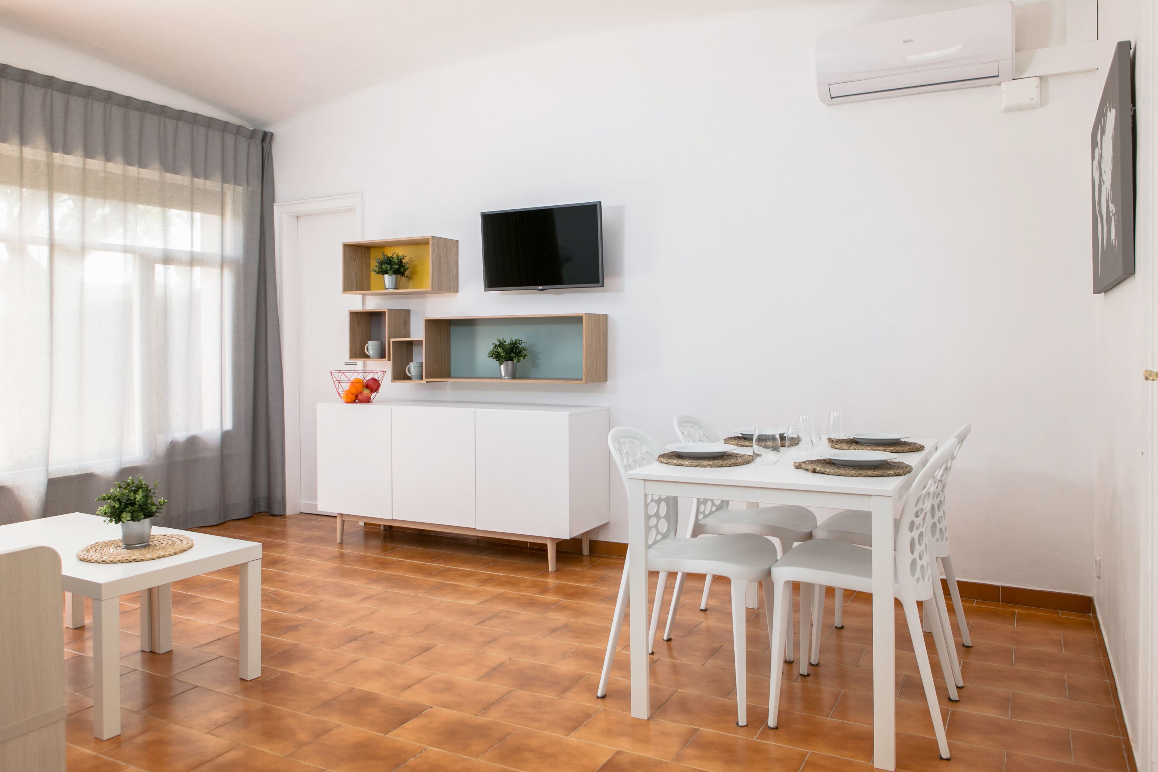 A modern, well-lit room with a dining table set for four, a white cabinet with shelves, a flat-screen TV mounted on the wall, and a window with sheer curtains.