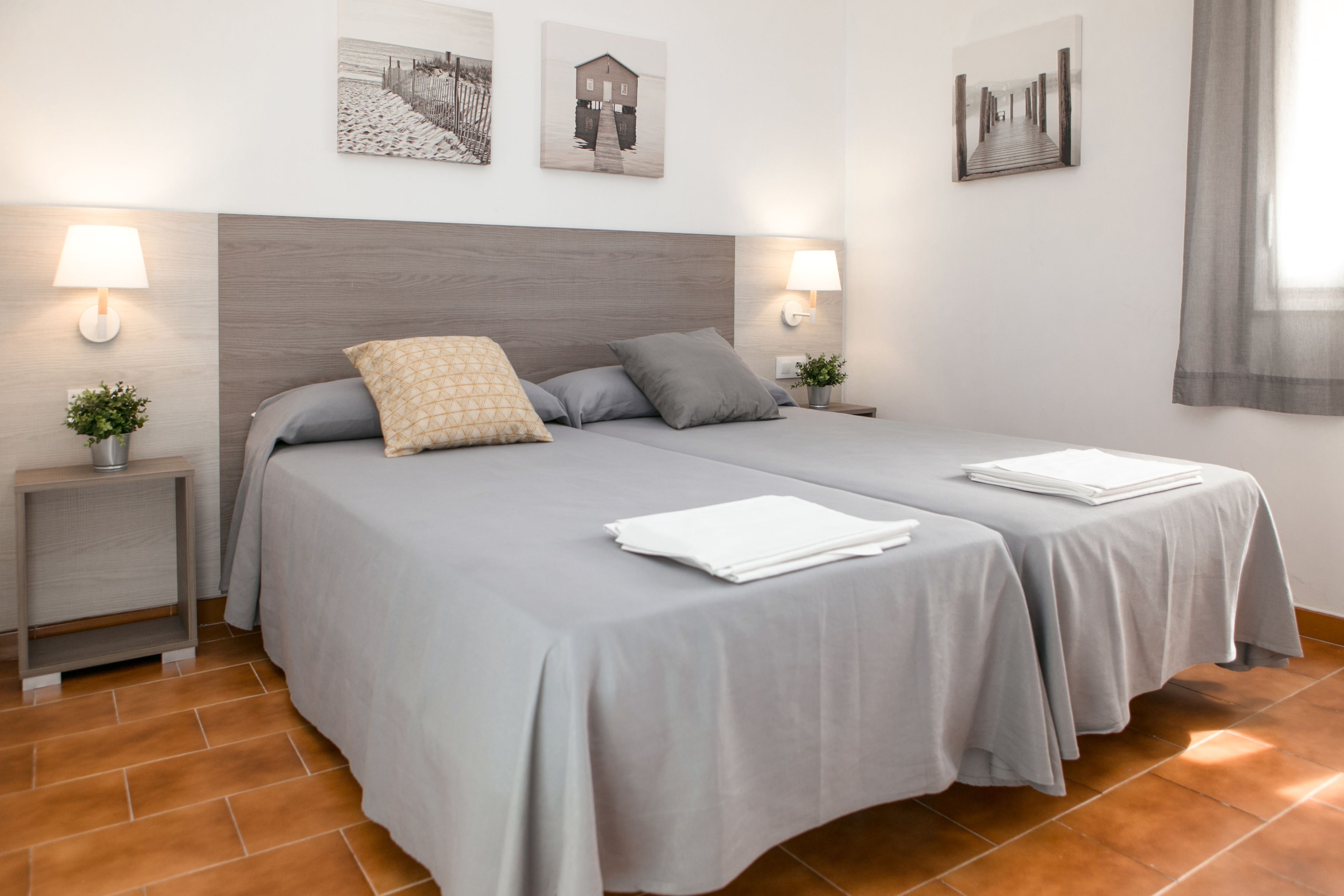 A double bed with grey linens, two bedside tables with lamps, and a wall with three framed pictures.