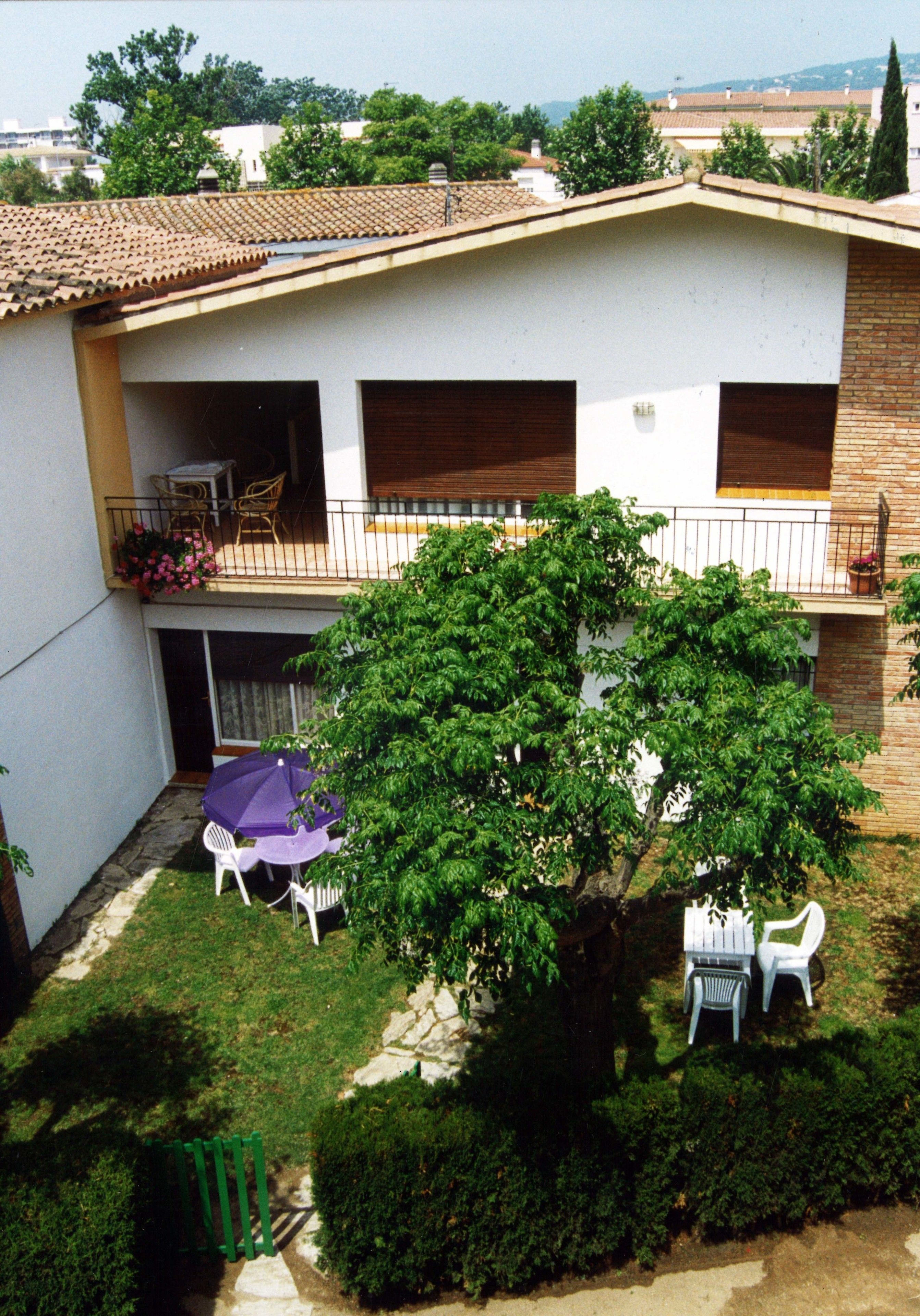 A residential building with a tiled roof, a balcony with a table and chairs, and a tree in the yard.