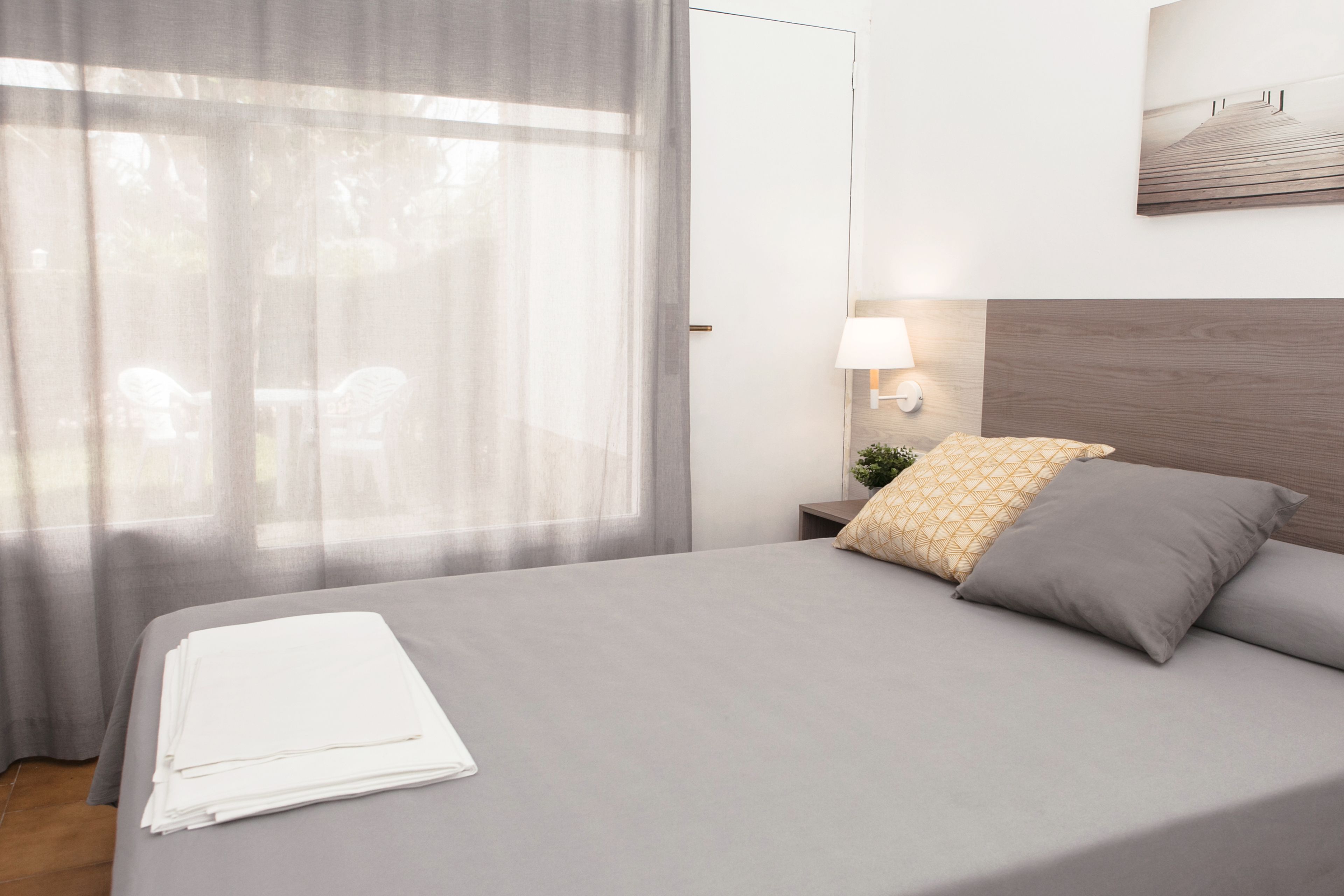 A neatly made bed with a grey bedspread and two pillows, a bedside table with a lamp, and a window with sheer curtains.