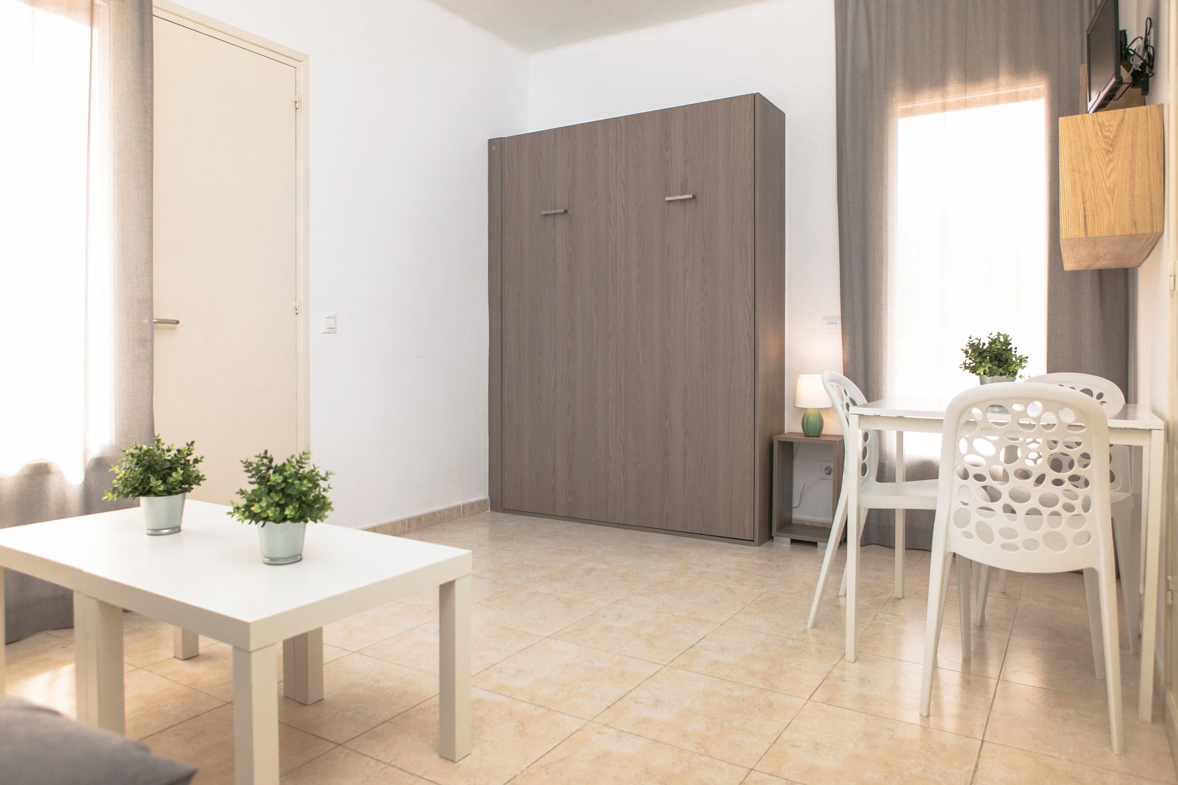 A modern room with a white table, two potted plants, a grey wardrobe, and a dining area with white chairs.