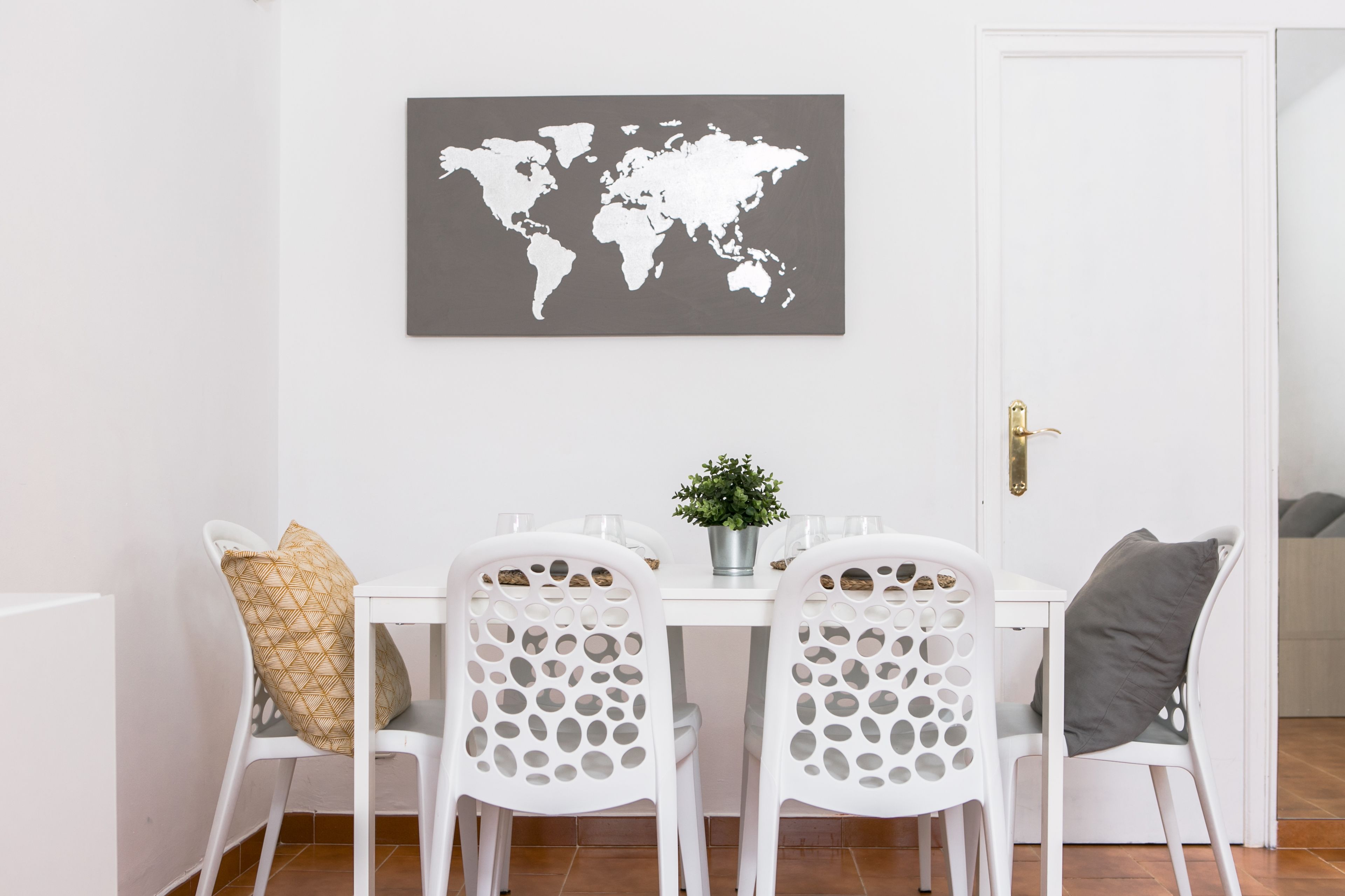 Un comedor con una mesa y sillas blancas, un mapa del mundo en la pared y una planta en maceta sobre la mesa.
