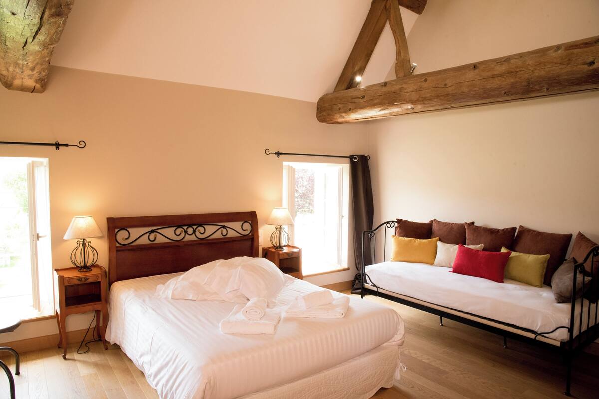 Quarto com cama grande, um sofá menor, duas mesas de cabeceira com luminárias e um vigamento de madeira no teto.