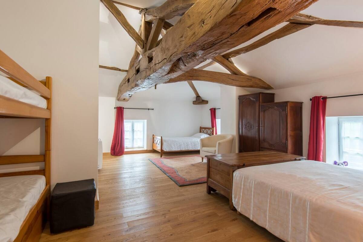 Um quarto amplo com teto alto, vigas de madeira, uma cama beliche, uma cama de solteiro, uma cadeira e um guarda-roupa.