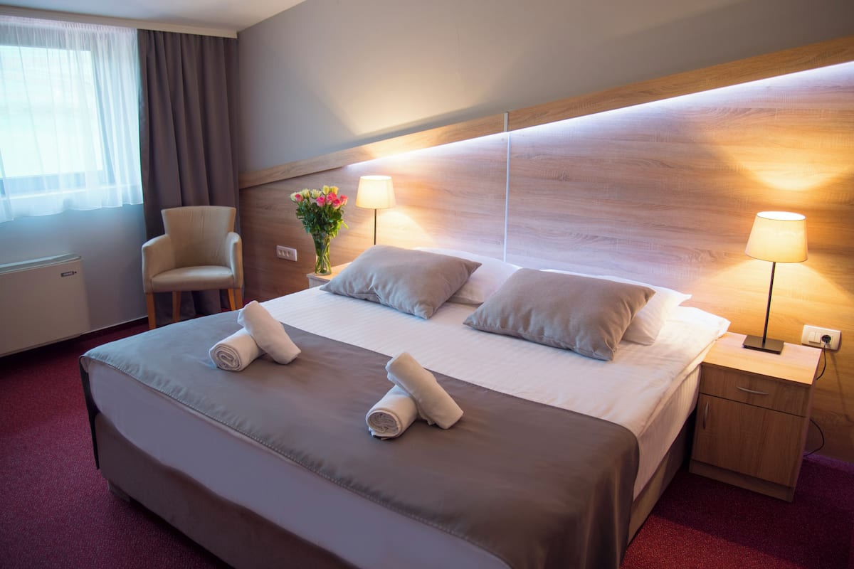 Una cama bien hecha con dos almohadas y dos toallas enrolladas, una lámpara de noche, una silla y un jarrón con flores sobre una mesita de madera.