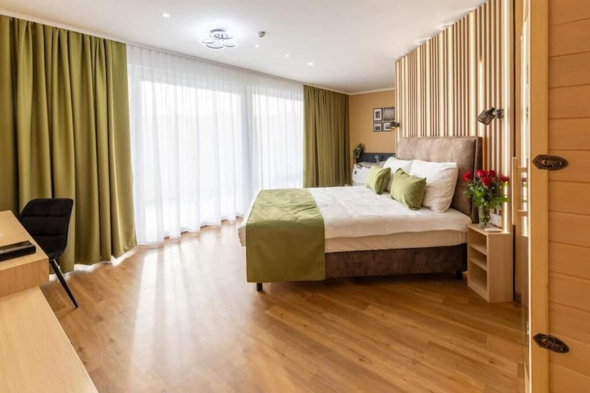 Habitación de hotel con una cama grande, un escritorio y una silla. Se ve una ventana con cortinas, una mesita de noche con un florero y una pared con listones de madera verticales.