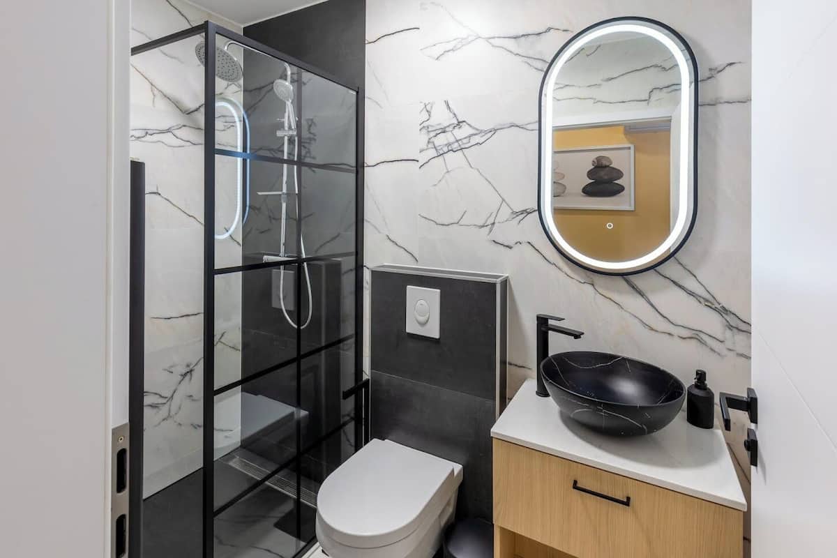 Un baño moderno con ducha de vidrio, inodoro, lavamanos de cerámica negra y espejo con luz.
