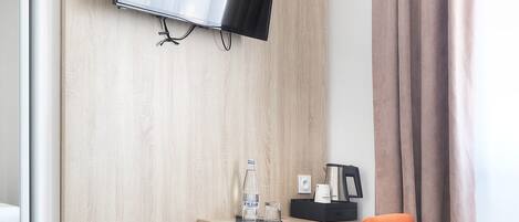 A hotel room with a wooden desk, a television mounted on the wall, an orange chair, a coffee maker, and a bottle of water.