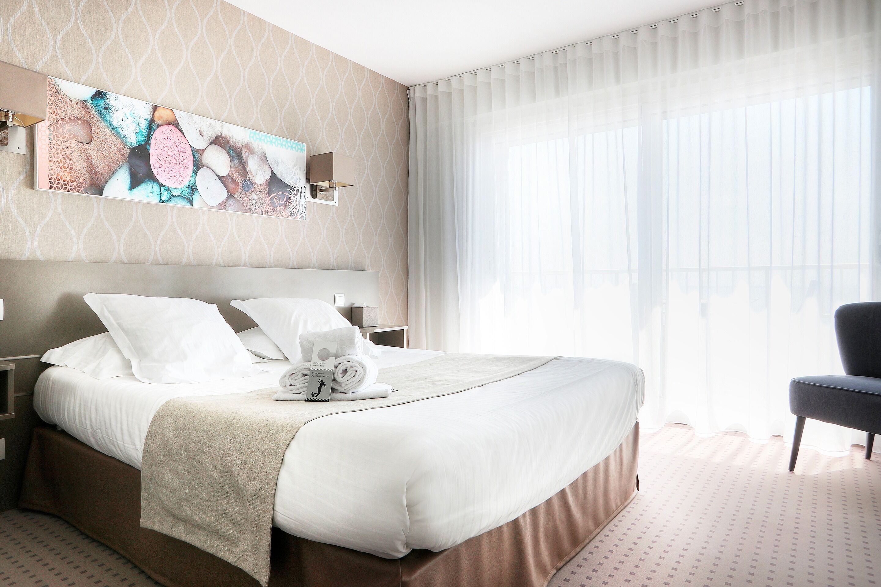 A neatly made bed with white linens and a beige throw blanket, a bedside table with a lamp, and a large abstract painting on the wall.