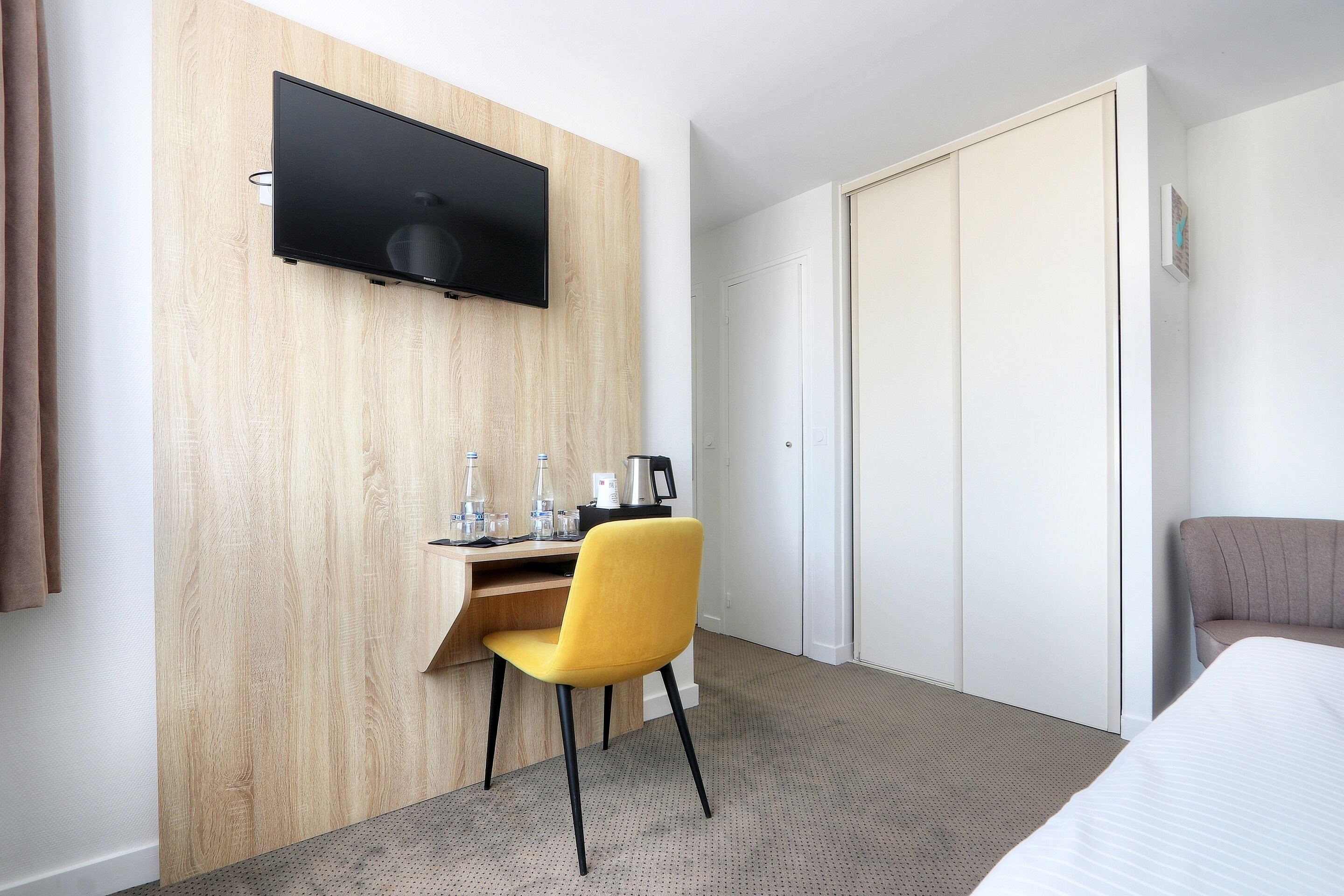 A hotel room with a wooden accent wall, a flat-screen TV mounted above a desk, a yellow chair, a bed, and a wardrobe.