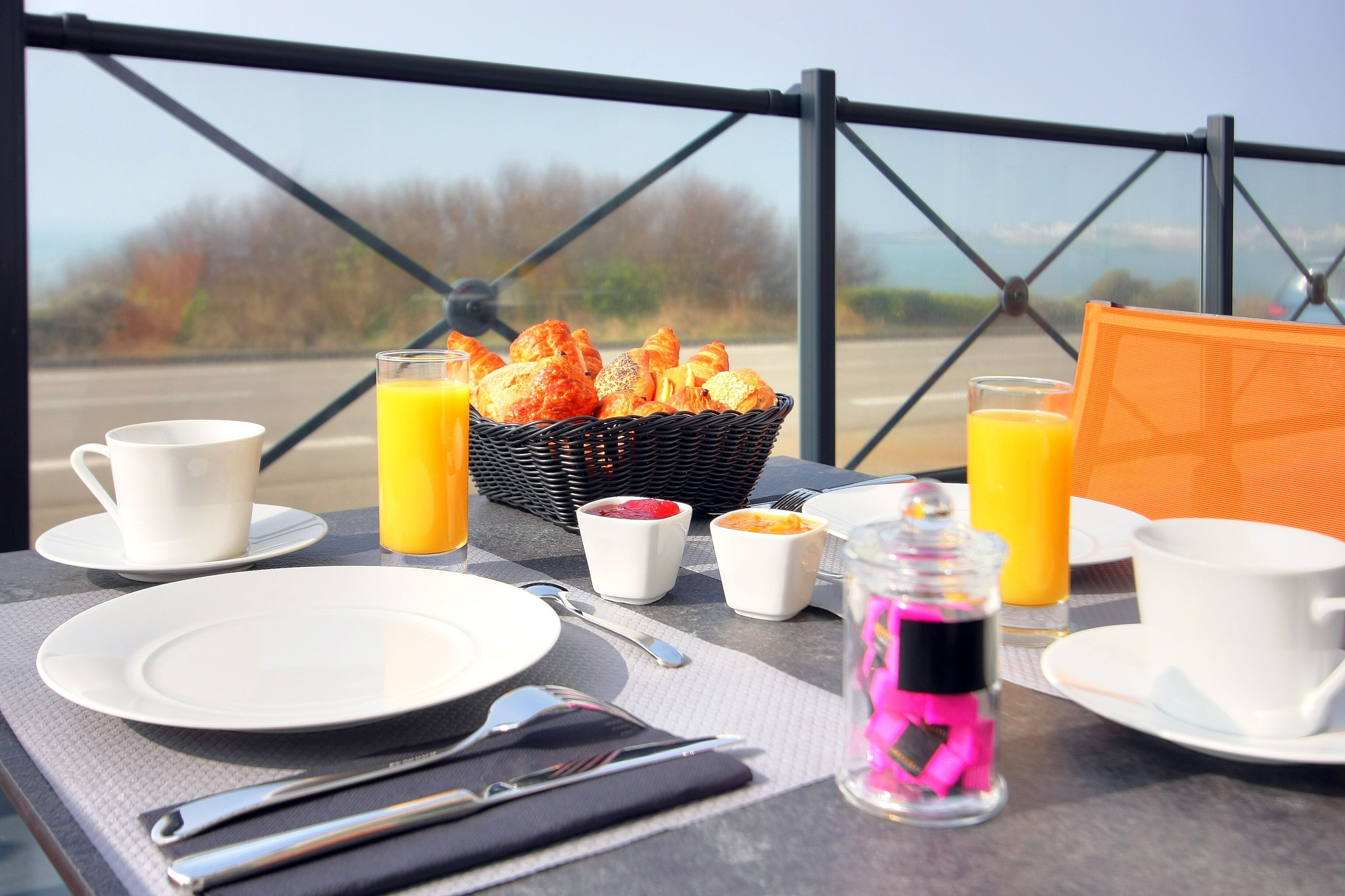 A breakfast setup with croissants, orange juice, and coffee on a table with a view of trees and a road.