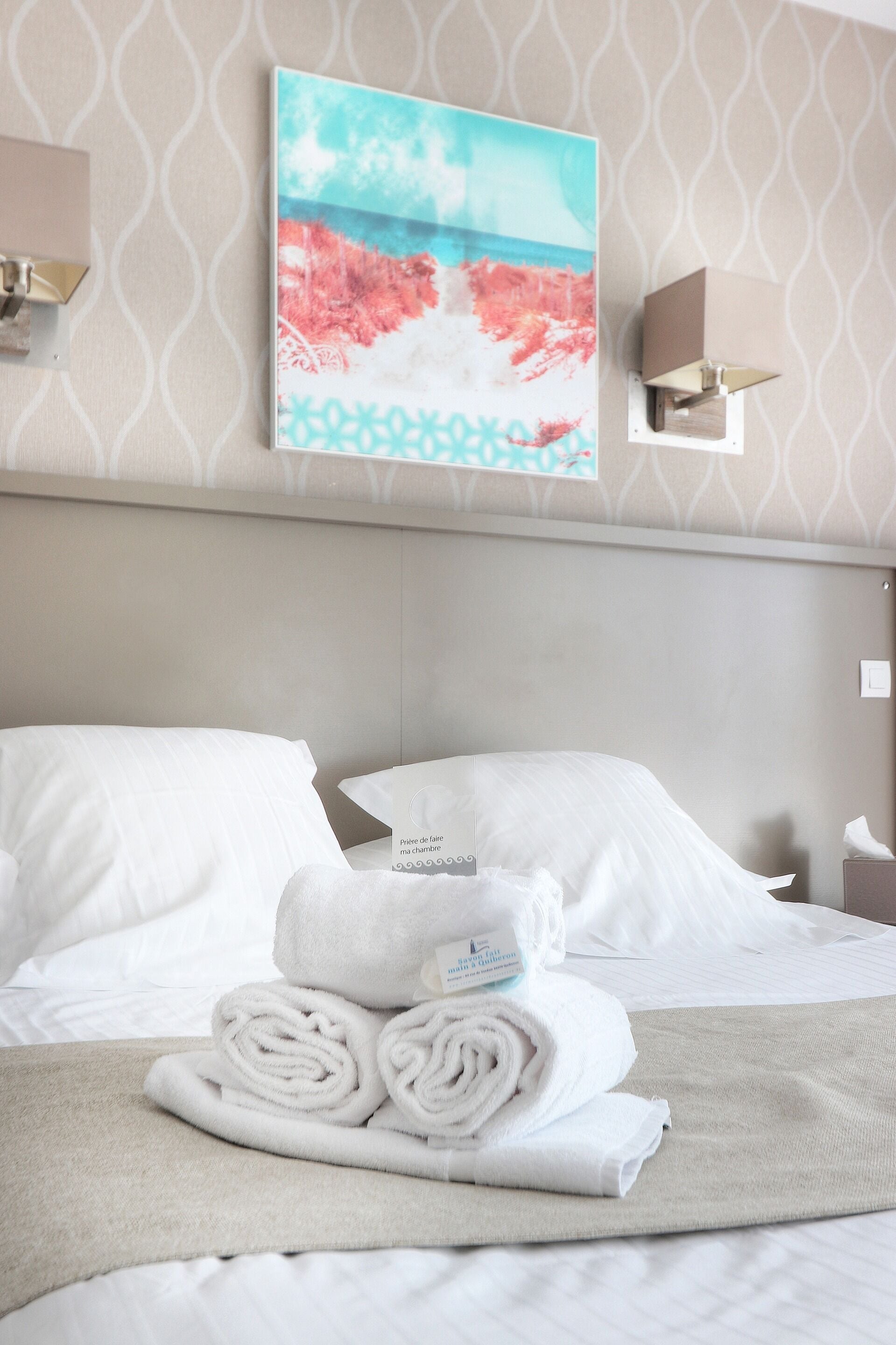 Un lit bien fait, avec des draps blancs et une serviette pliée posée sur une couvre-lit beige. Un tableau représentant une scène de plage est visible au mur, au-dessus du lit.