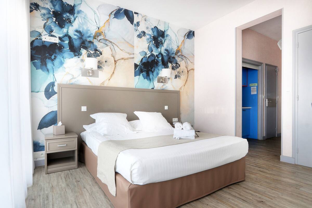 Ein modernes Hotelzimmer mit einem großen Bett, einem Nachttisch, einer Wandleuchte und einer Tür zu einem weiteren Raum.