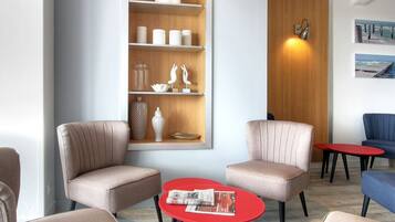 A modern waiting area with beige chairs, a red table, and a wooden shelf with decorative items.