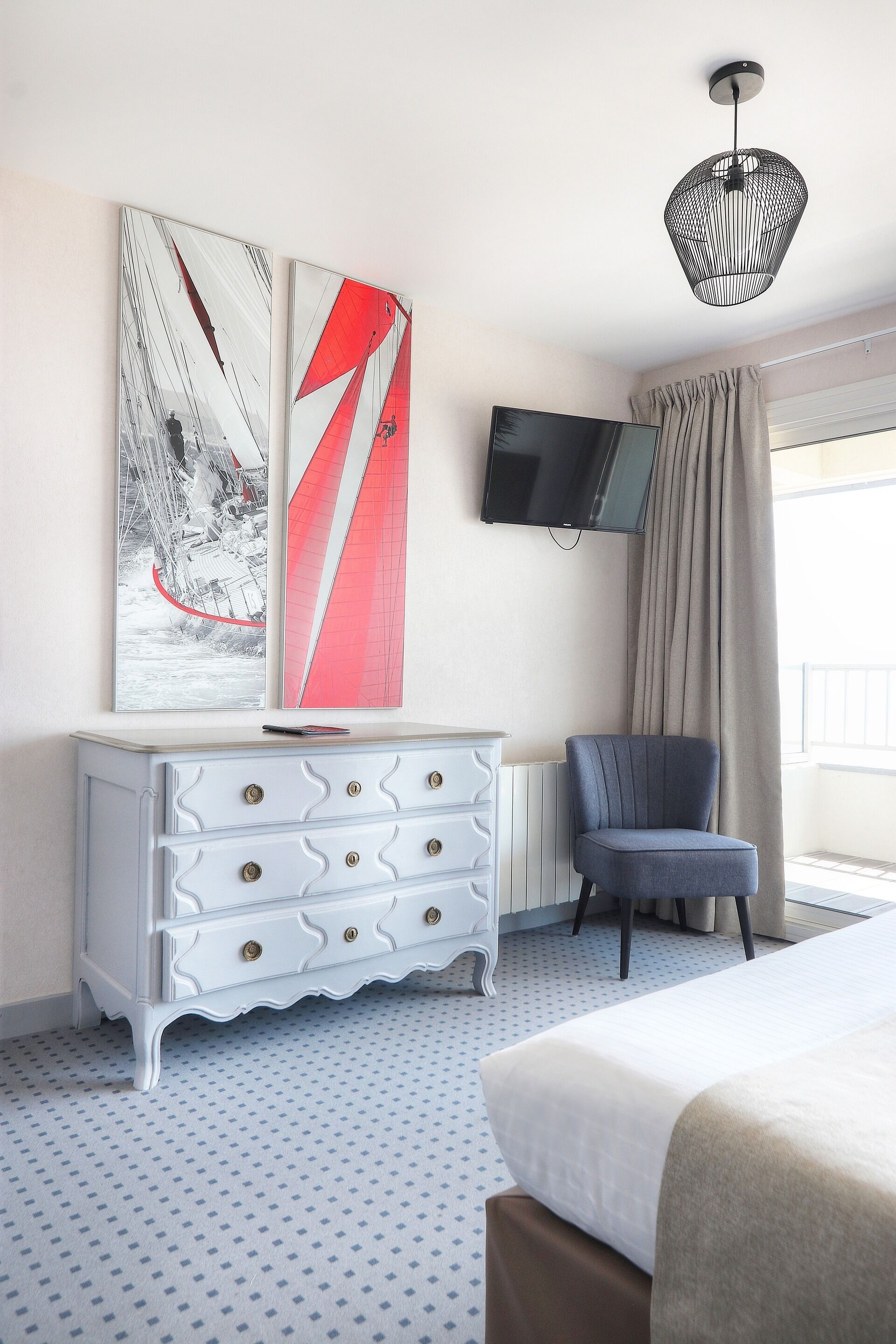 Habitación de hotel con una cama, un aparador blanco con cajones, un sillón azul, un cuadro abstracto de gran tamaño, un televisor de pantalla plana montado en la pared y una ventana con cortinas.