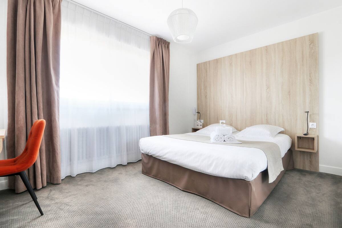 Ein Hotelzimmer mit Bett, Sessel, Nachttisch und einem großen Fenster mit Vorhängen.