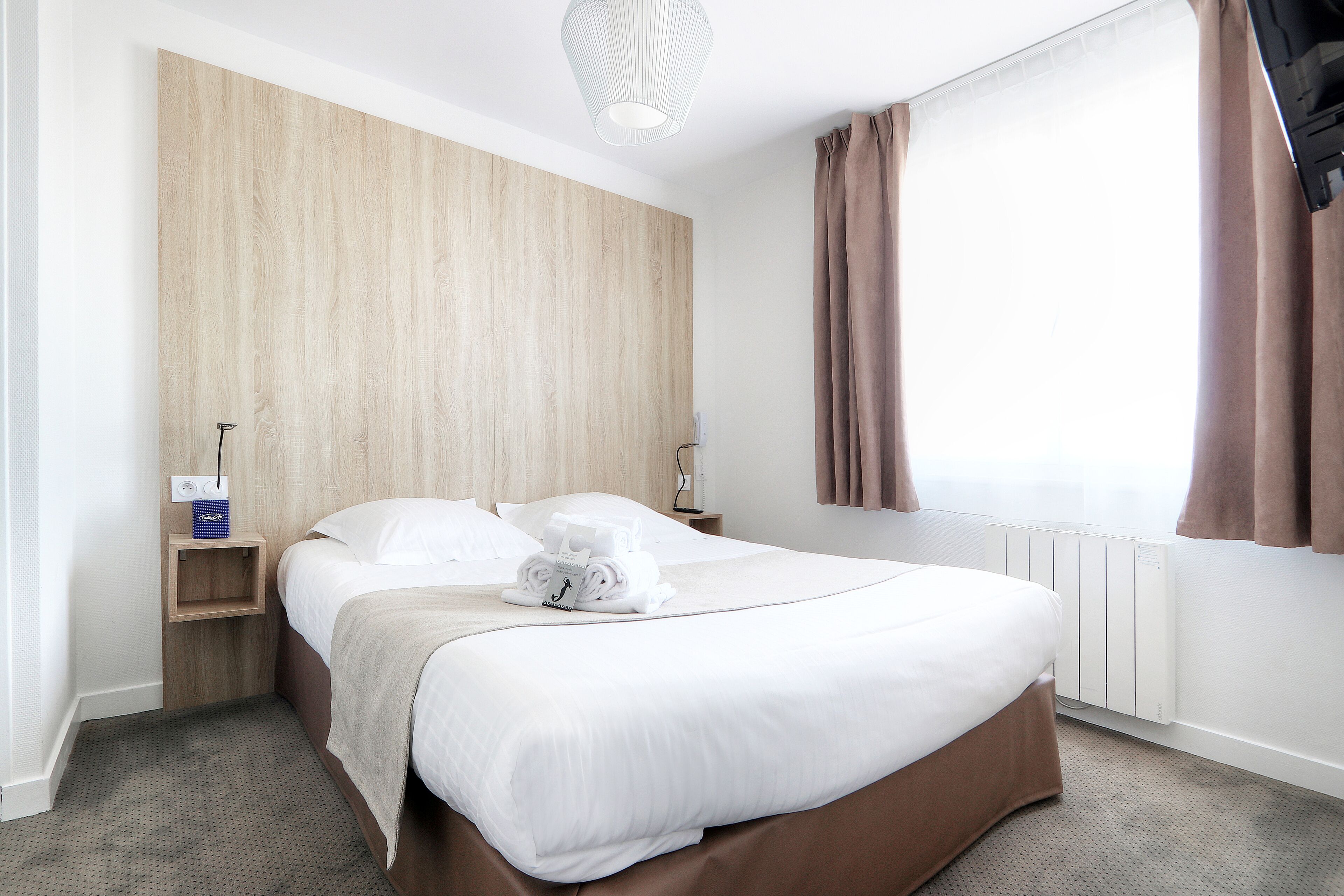 A neatly made bed with a brown bed skirt, a wooden headboard, a bedside table with a lamp, and a window with curtains.