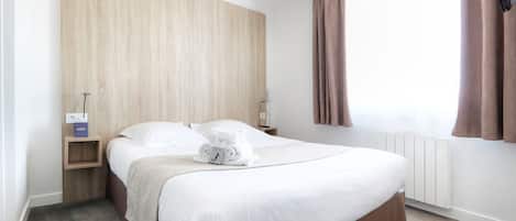 A neatly made bed with a brown bed skirt, a wooden headboard, a bedside table with a lamp, and a window with curtains.