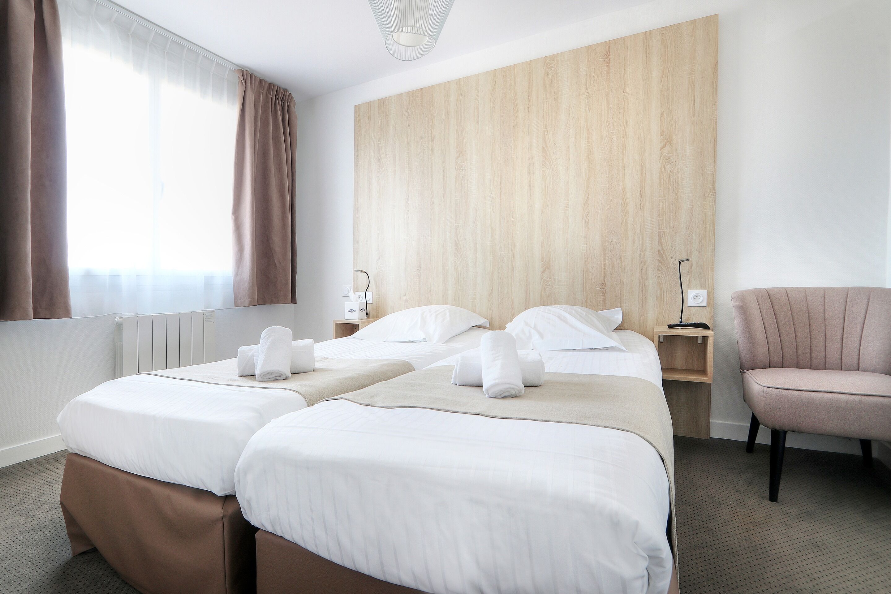 Habitación de hotel con dos camas, cabecera de madera, una silla, una mesita de noche y una ventana con cortinas.