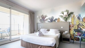 Een hotelkamer met een bed, nachtkastje, stoel, raam met uitzicht op buiten en een muur met een tropisch muurdecor.