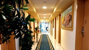 Pasillo de hotel con alfombra azul, plantas en macetas y un cuadro en la pared.