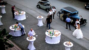 Mesas redondas al aire libre con comida y bebidas, rodeadas de gente y coches antiguos.