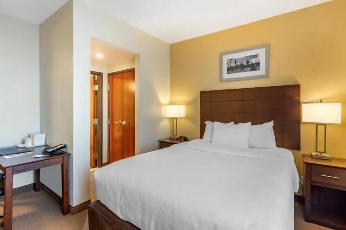 A hotel room with a large bed, two bedside lamps, a wooden headboard, a nightstand, a desk, and a framed picture on the wall.