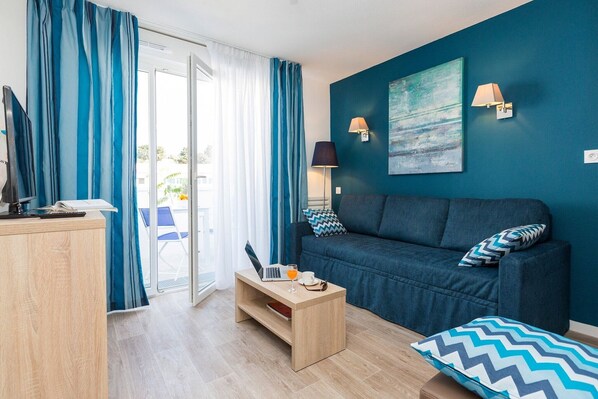 Una sala moderna con un sofá, una mesa de centro y un escritorio con una computadora. Se aprecia una vista al exterior a través de un ventanal.