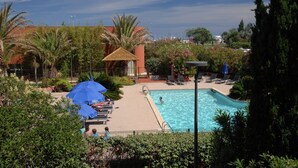 A swimming pool with lounge chairs and umbrellas, surrounded by greenery and a building with a red roof. - Coastal Retreat w/ Balcony (LE CAP D'AGDE)