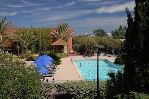 A swimming pool with lounge chairs and umbrellas, surrounded by greenery and a building with a red roof. - Coastal Retreat w/ Balcony (LE CAP D'AGDE)