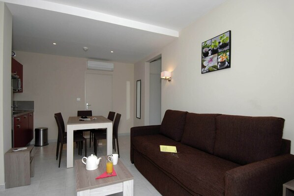 A living room with a brown sofa, a dining table with chairs, a kitchen area with a microwave, and a wall-mounted picture.