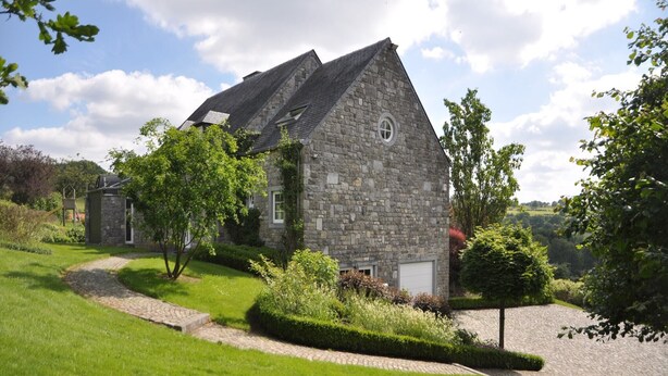 Een stenen huis met een schuin dak, omgeven door weelderige begroeiing en een kronkelend pad.