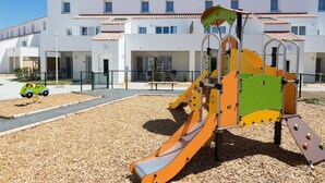 Ein Spielplatz mit Rutsche und Klettergerüsten, umgeben von einer Schotterfläche, mit einem Gebäude im Hintergrund.