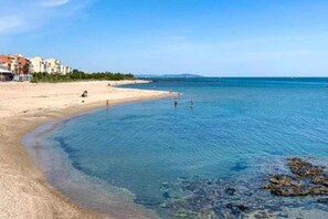 Ein Strand mit klarem Wasser, sandigem Ufer und Gebäuden im Hintergrund.