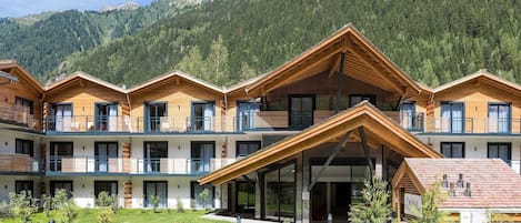 A modern hotel with multiple wooden balconies, a central entrance, and a mountainous backdrop.