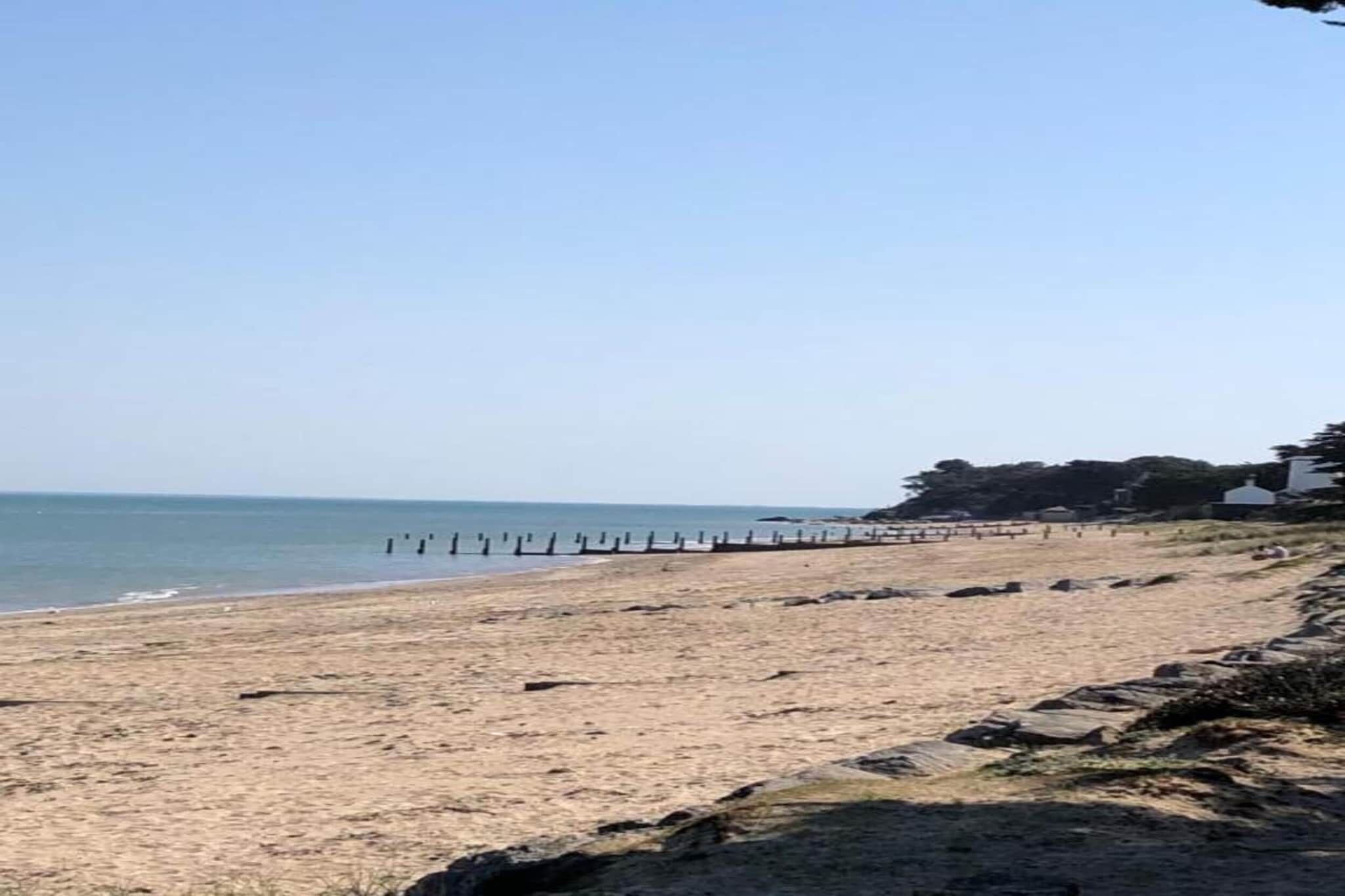 Une plage avec une jetée en bois qui s’avance dans l’eau, un ciel dégagé et une côte lointaine bordée de végétation.